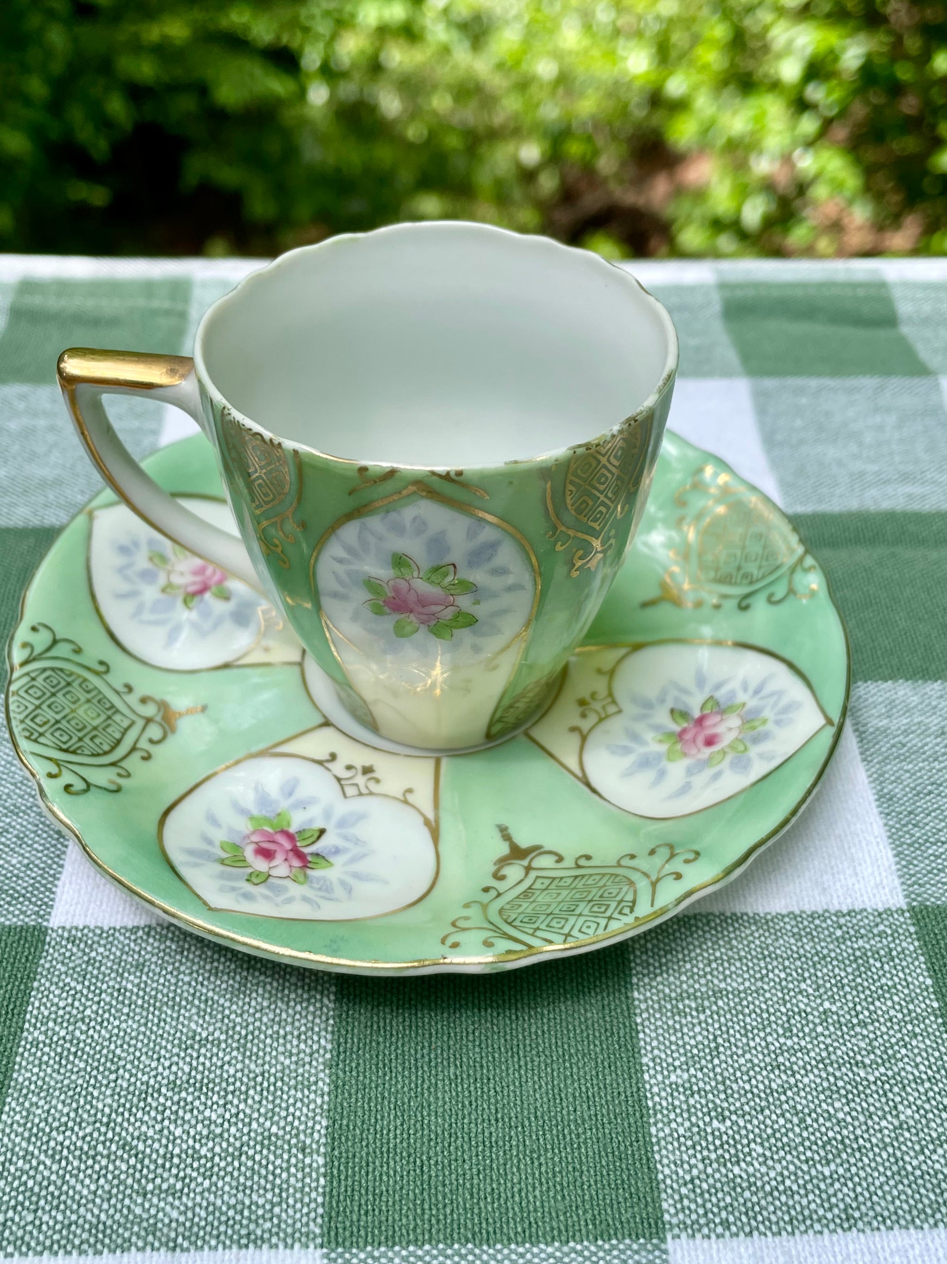 Green teacup and saucer with floral patterns on a green and white checkered tablecloth.