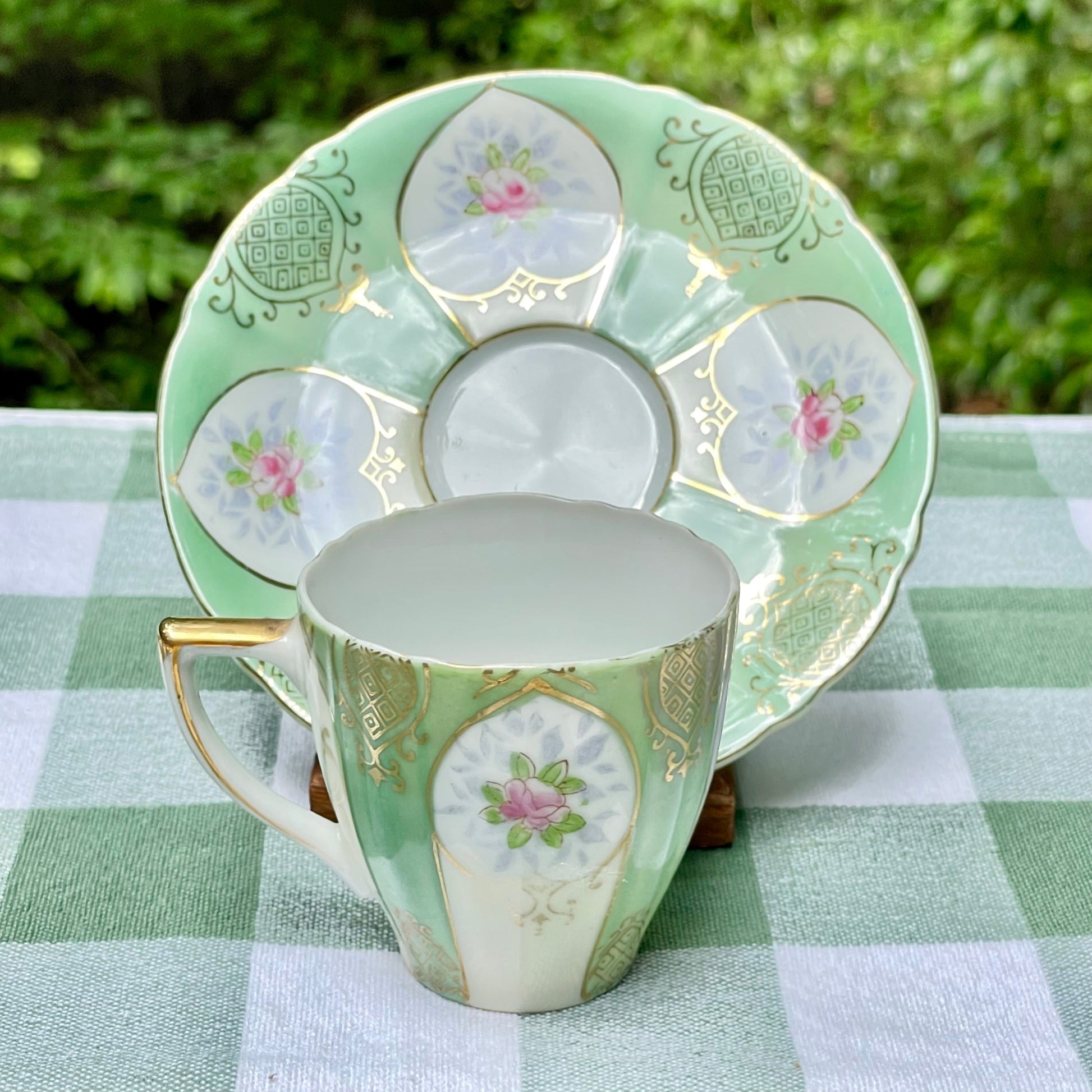 Decorative teacup and saucer with floral patterns on a green checkered tablecloth. thebrokenbirdcompany tea cup tea set green and gold pink rose buds UCAGCO Occupied Japan China tea cup