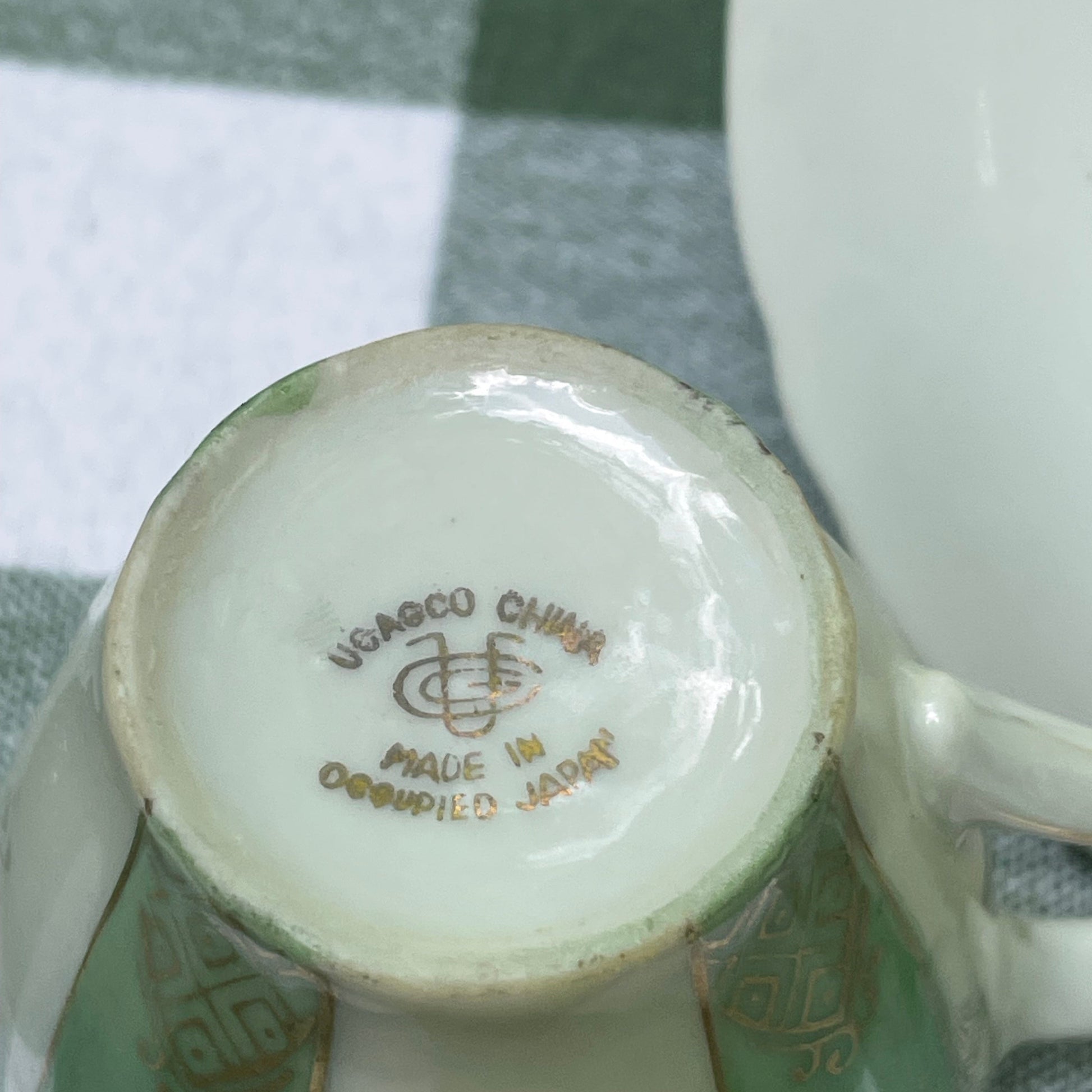 Close-up of a ceramic product with a visible brand logo on a checkered fabric background UCAGCO green and gold tea cup and saucer plate the broken bird company free shipping gift tea set
