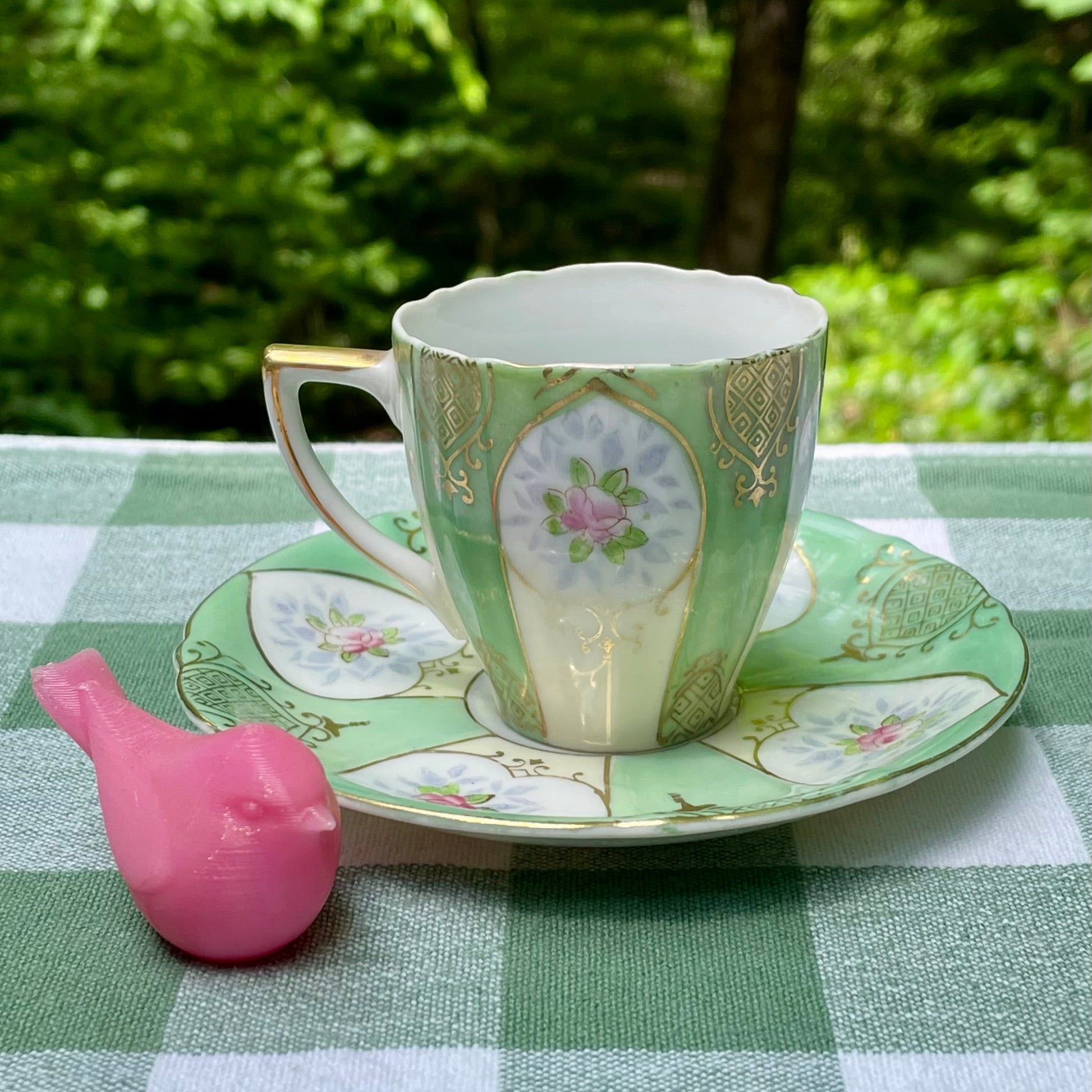 Teacup and saucer with floral design on a green checkered tablecloth outdoors.