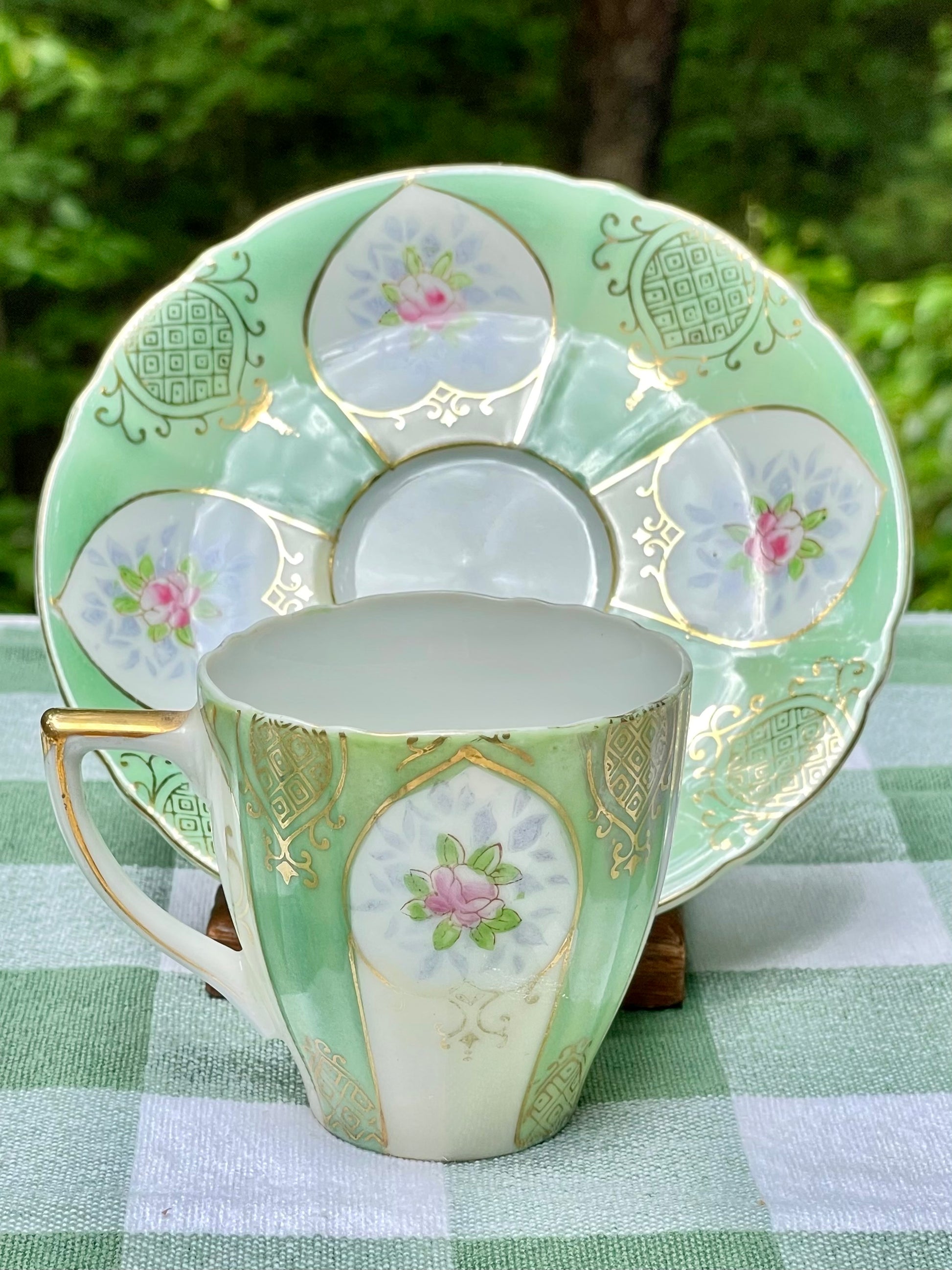 Decorative teacup and saucer set on a green checkered tablecloth with a blurred natural background