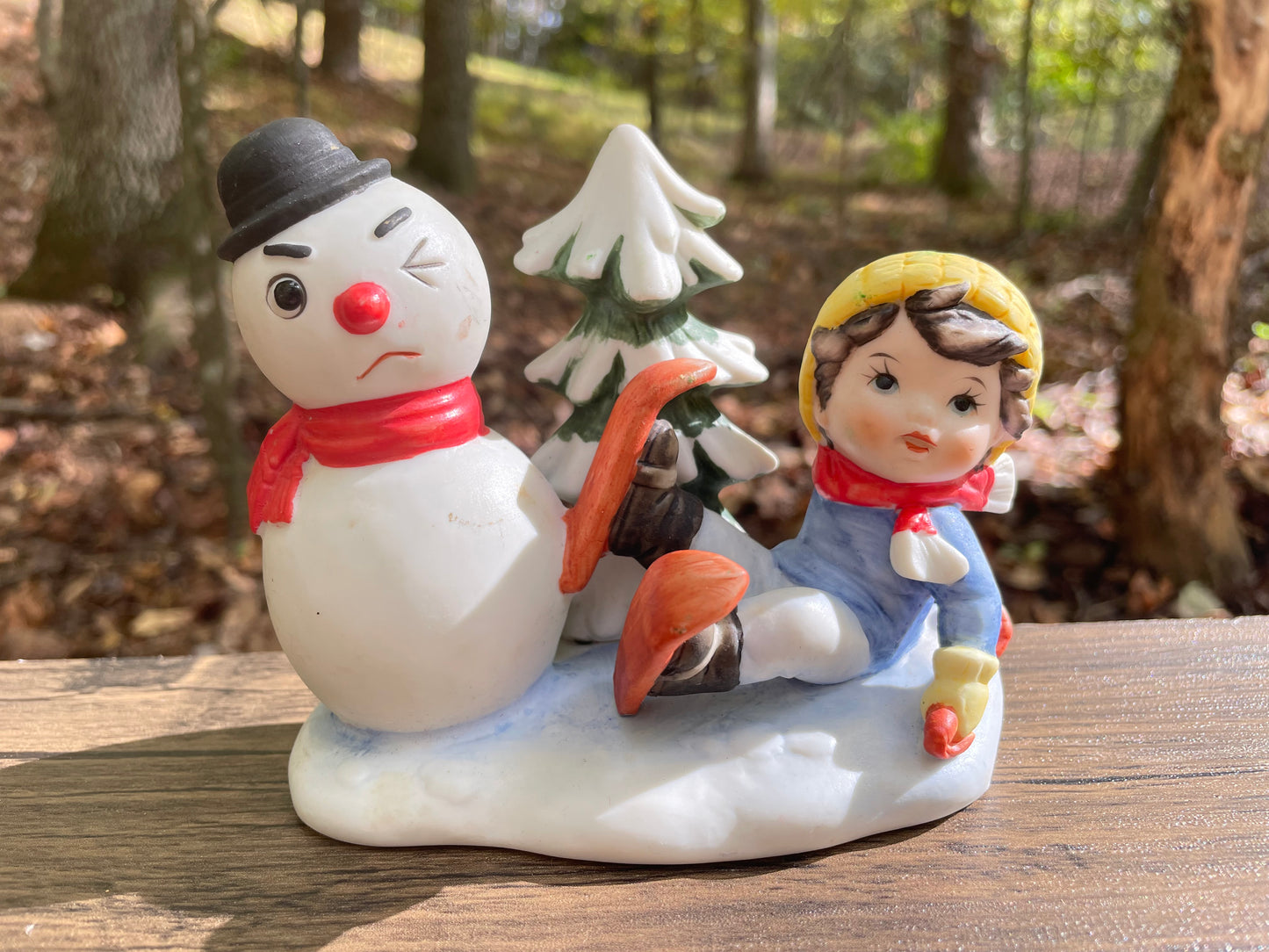 Vintage chirstmas cermaic figurine decor little boy with skis on sitting in the snow as if to slide into the snowman with one eye and winking with a christmas tree behind them around snow ground thebrokenbirdcompany vintage christmas decor ceramics gift 